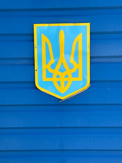 A blue and yellow shield-shaped sign, pointing to the right, is prominently mounted on a corrugated blue metal wall. The sign features the Ukrainian national emblem, the Tryzub (trident), depicted in bright yellow against a light blue background. Several small, dark screws or rivets are visible, securing the sign to the wall. The top right edge of the sign shows a slight bend or minor damage.
The background is a uniform, solid blue corrugated metal surface with distinct vertical ridges, suggesting it could be part of a fence, a shipping container, or a building facade. The lighting in the image is even and bright, indicating it was taken during daylight hours. There are no people visible, nor any specific activities taking place. The scene focuses solely on the emblem and its immediate surroundings. The presence of the Ukrainian national emblem in Balakliya, Ukraine, underscores the local national identity and resilience.