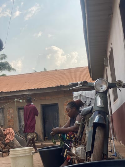 This image captures an outdoor domestic scene in the city of Aliade, Nigeria, under a light blue sky with scattered white clouds. In the midground, a woman with dark, reddish-brown hair tied up is seated on a low surface, wearing a dark sleeveless top and a patterned skirt. She is actively preparing leafy greens over a dark, rectangular basin positioned on the dirt ground in front of her. To the immediate right, the front portion of a vintage-style green motorcycle, featuring a large circular headlamp and handlebars, occupies the foreground, partially obscuring the woman. A white plastic bucket is visible on the ground in the lower-left foreground.
In the background, a second individual, wearing a maroon long-sleeved top and light-colored shorts or trousers, walks from right to left, facing away from the viewer. Behind this person, a black wheelbarrow is parked. The background features several simple residential buildings constructed with light-colored, possibly unpainted, walls and dark wooden doorways, topped with corrugated orange-red metal roofs. A clothesline strung between two structures holds various white and patterned laundry items. Palm tree fronds and other green foliage are visible above and to the left of the background buildings. The ground throughout the scene appears to be packed dirt or earth.