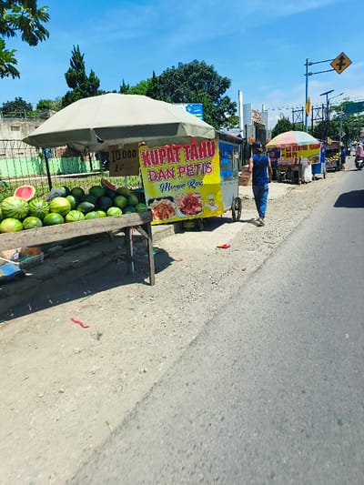 The scene takes place on a paved road in Margahayu, Indonesia, at the coordinates -6.985 latitude and 107.563 longitude. A man in a blue shirt and black pants walks on the right side of the road. He walks away from the camera, towards the right side of the image. Behind him is a food stall with a yellow sign that says "Kupat Tahu Dan Petis Mangga Rosa." The stall is selling watermelon and other items. Behind the stall is a green fence and a building with a green roof. The stall has a large white canopy that appears to be made of canvas. To the right of the man, there is another food stall. There is a large green tree at the beginning of the road to the left of the image and a blue sky above. The road is paved and grey with light patches of gravel. The road is lined with a ditch on the left side. The ditch contains small pieces of gravel, dry foliage, and a thin red string. The road is empty except for the man walking. The sky is mostly blue with white clouds.