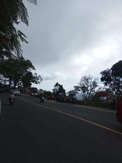 The image depicts an outdoor scene on a paved road overlooking a city, identified as Phoxay, Laos. The perspective is from the roadside, looking down a slight incline or curve to the right. The road surface is dark asphalt with a single solid yellow center line.

On the road, a white sedan is visible parked or moving slowly on the left side, near the curb, with additional vehicles partially obscured behind it. Two motorcycles are in motion: one in the foreground, traveling away from the viewer, and another further ahead, traveling in the same direction, with its rider wearing a yellow helmet.

To the left of the road, dark green foliage from trees extends into the upper left quadrant, creating a canopy effect. Along the right side of the road, a low, light-colored stone wall delineates the edge, beyond which the landscape drops off, revealing a distant cityscape or town with scattered buildings.

Several informal vendor stalls are set up along the right shoulder of the road, next to the wall. One prominent stall features a striped umbrella (white, grey, light blue) and a metal cart underneath. Another stall further right has a red umbrella. People are visible near these stalls and along the roadside, some appearing to be vendors or customers. Parked motorcycles are also present near the stalls.

The sky is heavily overcast with significant cloud cover, exhibiting various shades of grey and white, with brighter patches indicating obscured sunlight. The overall lighting is diffuse.