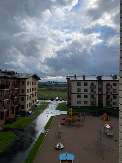 The image depicts an aerial view of a residential courtyard in Poselok Kornevo, Russia. The scene features two multi-story apartment buildings, one clad in light brown brick and the other in dark brown brick. Both buildings have numerous windows and balconies, suggesting a relatively modern construction.
Adjacent to the buildings is a playground area with sand surfacing. Visible playground equipment includes a red and yellow climbing structure with slides, a small red-roofed playhouse, and a net climbing structure. A small carousel is also present. A paved walkway, wet and reflecting the overcast sky, winds through the courtyard, bordered by patches of green grass. Several cars are parked along the walkway, and a few are visible in the distance.
The sky is filled with dramatic, heavy clouds, indicating potentially rainy or recently rained weather. The overall lighting suggests it is daytime, possibly late afternoon or early evening, due to the diffused light and the dark, wet surfaces. The mood is somewhat somber due to the cloudy weather and the wet ground. There are no people visible in the image. The setting appears to be a suburban or semi-urban residential area. No text is discernible in the image.