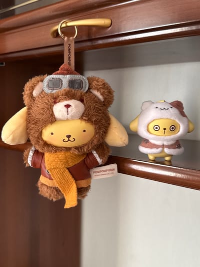 Two Pompompurin plush toys are in the image. One plush is brown and dressed as a pilot, complete with a scarf and goggles, attached to a gold keychain with the text "POMPOMPUP." The other plush is white and brown, styled as a winter character. The toys are resting against a wooden surface. The location is not identifiable as Severnyi, Russia, based on the visual information provided.