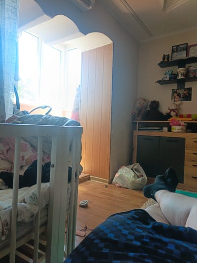 This image depicts a bedroom scene in Pivdenne, Ukraine. The room is bright, with sunlight streaming through a large window, suggesting it is daytime. The setting appears to be a child's bedroom or a room with baby items.
In the foreground, a person's legs, clad in dark socks, are visible, resting on what appears to be a light-colored rug or blanket. The person is wearing dark blue checkered pajama bottoms. To the left of the frame, a white crib is partially visible, filled with blankets and a stuffed animal. The crib is positioned near the window, which has a light blue curtain on the left.
The background of the room shows a wooden door on the right side and a tall, dark dresser with wooden drawers. On top of the dresser, there are several shelves displaying various items, including teddy bears, framed pictures, and what looks like a trophy or certificate. A large white plastic bag lies on the wooden floor near the dresser.
The overall impression is one of a lived-in, cozy space, possibly a nursery or a child's bedroom, bathed in natural light. There is no discernible text in the image.