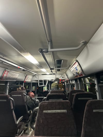 The interior of a bus is depicted. Passengers are seated in brown upholstered seats with silver accents. A white wall with silver railings runs along one side. Overhead compartments are visible. The bus appears to be in motion as lights can be seen through the windows at night. There is a mix of empty and occupied seats.