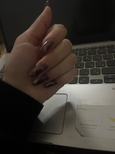 The image captures a close-up, overhead view of a person's hand resting on a silver laptop. The scene is set indoors, likely at a desk or table, with the overall lighting appearing somewhat dim, casting shadows on the laptop's surface. Given the provided context, the location is Jaramana, Syria.

The hand features long, manicured nails, which are painted in a dark, possibly marbled or shimmering purple-brown hue. Most of the nails exhibit a coffin or tapered square shape, while the pinky finger's nail appears slightly more pointed. The hand is positioned over the laptop's keyboard and touchpad area, suggesting an interaction with or proximity to the device.

The laptop is a silver ASUS model, as indicated by a promotional sticker on its palm rest. Visible text on this sticker includes "ASUS Sptop" (likely "ASUS Laptop"), "NanoEdge Display," "Immersive visuals," "Light," and "Extremes," alongside graphic elements such as a laptop screen icon and a lightbulb. The keyboard prominently displays both English and Arabic characters, with examples like "ctrl," "ز," "ظ," "ل," "ک," and "ط" clearly visible. No specific time of day or emotional state can be discerned from the image.