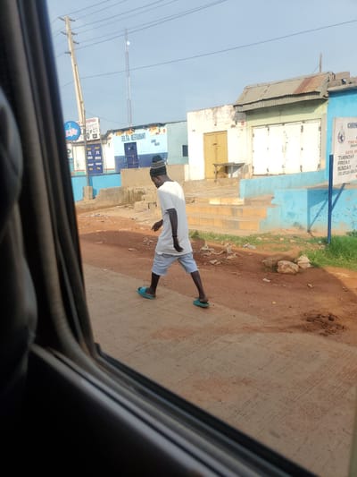 This is a photograph taken from inside a vehicle, looking out at a street scene in Goni Gara, Nigeria. The foreground is dominated by the dark interior of the vehicle's window frame. In the mid-ground, a person wearing a white t-shirt, light blue shorts, a dark beanie, and blue flip-flops is walking from left to right across the image. They are walking on a paved surface that appears to be a sidewalk or a part of the road. Behind the person, there are buildings with various paint colors and architectural styles, including some with corrugated metal roofs. One building has a sign that reads "DELTA RESTAURANT" and "AMB. BAR". Another sign, partially visible, displays text related to "CHRIST THE KING" and lists days of the week. The environment appears to be somewhat dusty and shows signs of wear and tear, with some debris scattered on the ground. Utility poles and wires are visible in the background, indicating the presence of infrastructure. The sky is clear and pale blue.