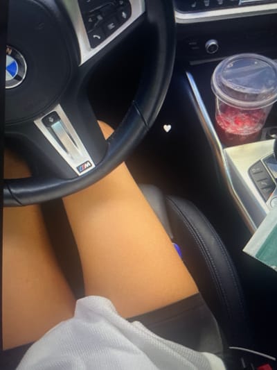 The image shows a close-up view of the interior of a car, specifically the steering wheel and the area around the center console. A person's legs, covered by shorts and partially by a white garment, are visible in the foreground. On the steering wheel, a BMW logo is clearly discernible. To the right of the steering wheel, a clear plastic cup filled with a red liquid, possibly a beverage with fruit, is placed in a cup holder. The center console area has buttons and a dial. The overall scene suggests the person is a passenger or driver in a modern car, and the image was likely taken from a personal device. There is no information available to determine the specific location within the city of Kołobrzeg, Poland.