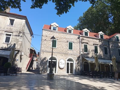 The image shows a street in the city of Trebinje, Bosnia and Herzegovina, on a sunny day. The scene is dominated by traditional stone buildings with red tiled roofs and green shutters. In the foreground, there is a paved plaza with several outdoor seating areas for cafes. A prominent lamppost stands in the center of the plaza.
One building, on the right side of the frame, features a storefront with arched windows and a sign that reads "dior BEAUTY & LIFESTYLE". Adjacent to it, another establishment, "caffe Natale", is visible with tables and chairs set up outside under awnings.
To the left, another building, with a more weathered facade, has multiple windows and an external staircase leading to a higher level. Large umbrellas are set up outside one of the establishments on the left. Lush green trees with visible branches and leaves overhang the scene, casting shadows on the plaza. The sky is clear blue. There are no people clearly visible in the image.