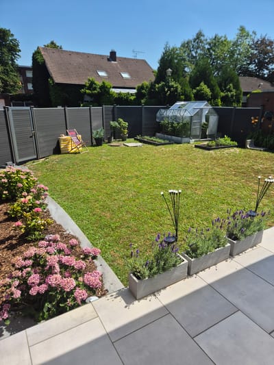 This is a medium-shot, eye-level, outdoor, daytime photograph taken in the city of Lüdinghausen, Germany. The scene depicts a backyard with a neatly mowed green lawn. In the foreground, a modern patio area with large, light-colored square tiles is visible. Along the edge of the patio, three rectangular planters contain lavender plants. Next to these planters, two solar-powered garden lights with multiple white LED bulbs on thin black stems are positioned.

To the left of the lawn, a bed of pink hydrangeas is mulched with wood chips and bordered by a concrete strip. Behind the hydrangeas, a dark gray slatted fence encloses the yard. A striped yellow and red lounge chair leans against the fence near a small stack of items, including what appears to be a yellow tarp.

Further into the yard, a small greenhouse with a clear plastic roof and white frame is visible. Inside the greenhouse, plants are growing. In front of the greenhouse, there are raised garden beds with dark frames, also containing plants. Scattered around the lawn are other small plants and garden elements, including a light pole. The background features houses with tiled roofs and mature trees, under a clear blue sky. The overall impression is of a well-maintained residential garden.