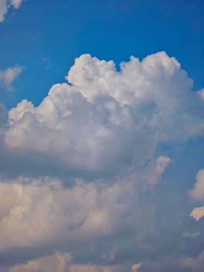 The image is a close-up of a bright blue sky filled with voluminous white and gray clouds. The clouds are varied in shape and texture, with some appearing fluffy and illuminated by the sun, while others are darker and more dense. The lighting suggests it is daytime, possibly late afternoon, given the warm tones present in some of the clouds. There are no discernible objects, people, or structures visible in the frame, and the image solely focuses on the atmospheric conditions. The location of Pogodaevo, Russia, is not ascertainable from the visual information provided in the image.