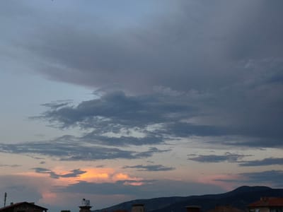The image captures a panoramic view of a cloudy sky at dusk over the mountainous landscape of Krichim, Bulgaria, with a few buildings visible in the foreground. The sky is largely overcast, dominated by heavy, dark grey clouds in the upper and middle sections, suggesting a dense cloud cover. Below these, lighter grey and wispy clouds are interspersed across the mid-ground.

Towards the bottom of the sky, a striking sunset is taking place, with vibrant patches of orange and soft pink light illuminating the clouds and the horizon. This warm glow contrasts sharply with the cooler, darker tones of the clouds above.

In the lower portion of the image, the dark silhouettes of mountains are visible on the horizon, stretching across the middle-right. In the immediate foreground, several structures are partially visible. On the far left, a section of a building with a reddish-brown tiled roof and an antenna or mast is present. To its right, what appears to be a utility structure or water tower with a light-colored top stands out. Further right, other reddish-brown tiled roofs and chimneys are scattered, and in the bottom right corner, a segment of another building with a red tiled roof and a small white-framed window is seen.

The scene depicts the transition from day to night, characterized by the dramatic interplay of heavy clouds and the warm hues of the setting sun, offering a moody yet beautiful natural spectacle over a typical settlement in the region. There is no visible text in the image.