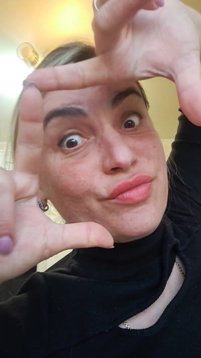 A woman with fair skin, dark eyebrows, and brown eyes is positioned at an unusual angle, seemingly upside down in the image. Her lips are red. She's wearing a black turtleneck sweater with a gold chain visible near the neckline. She is making a framing gesture with both hands, with her fingers close to the camera. Her fingernails are painted with a light purple polish. A silver ring is visible on one of her fingers. There appears to be a lamp with a cylindrical shade visible in the lower left corner of the image. The background is a light beige or yellow color. The location in the city of Меткомелино, Russia, is not discernible from the image.