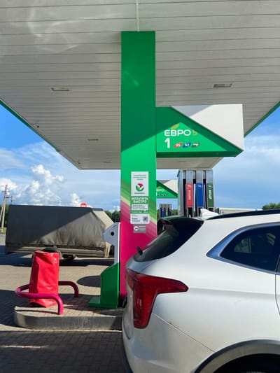 The image depicts a gas station canopy with a white car parked underneath. The canopy is supported by a large green pillar that extends upwards. Above the car, a sign reads "ЕВРО 5" and lists fuel grades 95 and 92. There are also fuel pumps visible. To the left of the pillar, a large red object, possibly a bag or a cushion, is supported by a pink metal structure. In the background, a large, tarp-covered vehicle is parked. The ground is paved with interlocking bricks. The sky is blue with scattered clouds. The scene appears to be a bright, sunny day at a gas station. The location is identified as a gas station in Arkhangelskoye, Russia.