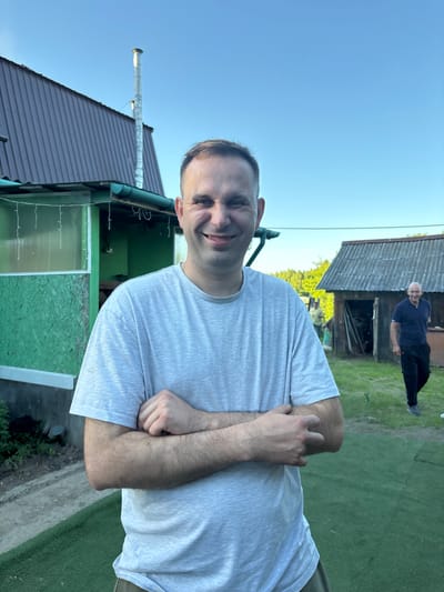 This image, taken in Urmanets, Russia, depicts an outdoor scene with three adult males and two small buildings under a clear blue sky.

In the foreground, a light-skinned adult male is centrally positioned, smiling broadly and looking directly at the viewer. He has short, dark hair with a receding hairline. He is wearing a light grey short-sleeved t-shirt and olive-colored trousers, with his arms crossed over his chest.

Behind the main subject, to his left, is a small, single-story building with light green walls and a dark brown corrugated metal roof. A green gutter system is installed below the roofline, from which white string lights or decorations hang. A tall, slender, shiny metal chimney extends vertically from the roof, rising prominently against the clear sky.

In the mid-ground, to the right of the main subject, a second light-skinned adult male is visible, walking from right to left. He has grey hair and is wearing a dark blue polo shirt and dark trousers. He appears to be looking generally forward or slightly towards the main subject. Behind him is a rustic, weathered wooden shed with a dark, possibly slate or shingle, pitched roof. An open doorway reveals a dark interior with some indistinct objects.

A third individual, partially obscured by the main subject and the shed, is visible in the background-left, wearing a light-colored shirt.

The ground in the foreground and mid-ground is covered with green artificial turf or a well-maintained lawn. Natural green foliage, including trees, is visible in the distant background, particularly on the left. The sky is uniformly clear and bright blue, indicating fair weather and daylight conditions. A thin wire or string extends horizontally across the mid-ground from the green building.