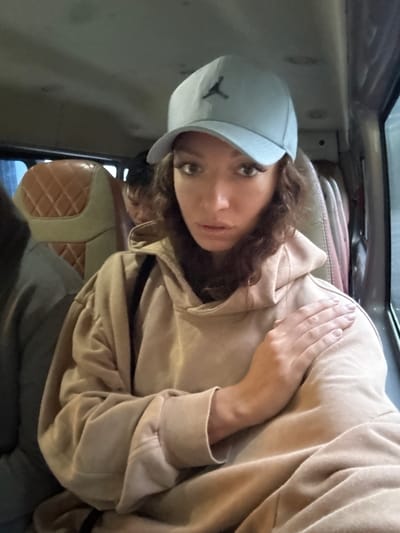 A selfie taken indoors, likely in a vehicle, shows a person with brown hair wearing a light blue baseball cap with a black logo and a beige hooded sweatshirt. The person's right hand is partially visible near their face. The interior of the vehicle includes diamond-patterned seating and a window visible in the upper left corner. No specific details suggest the exact location in Binh Lu, Vietnam.