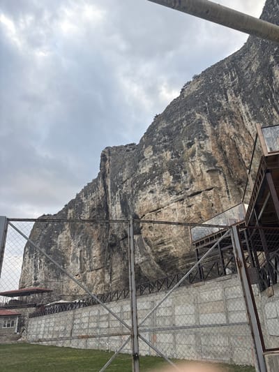 The image displays a prominent, rugged cliff face composed of varied grey, brown, and tan rock formations, rising steeply under a densely overcast, light grey sky. The scene is located in Bakhchysarai, Ukraine. In the foreground, a chain-link fence with vertical metal posts extends across the lower and middle sections of the frame. Behind this fence, a grey concrete retaining wall with a square block pattern is visible, topped by a dark metal railing. To the far left, behind the fence and wall, a building with a red-brown pitched roof and lighter-colored walls is partially visible, with another similar roofline further in the background. To the right, a multi-level structure constructed of dark metal framework and transparent glass panels is built into the base of the cliff, featuring visible stairs and platforms. A thick, light-colored cylindrical object or beam diagonally crosses the upper right corner of the frame, partially obscuring the cliff and sky. A patch of green grass is present in the lower-left corner of the foreground. No individuals or active interactions are depicted.