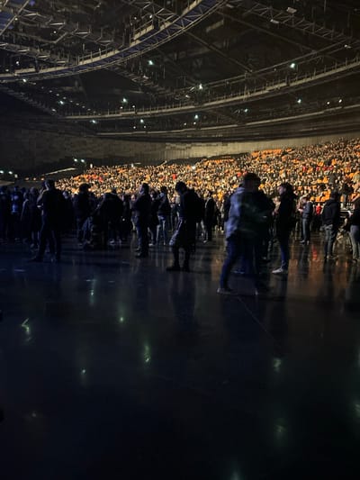 An arena filled with many people. The arena's metal framework is visible on the left side. The crowd is tightly packed and stretches along the left side of the image. In front of the crowd, a darker area reflects some of the lights. It is unclear exactly where the people are located, but it is likely near a stage.