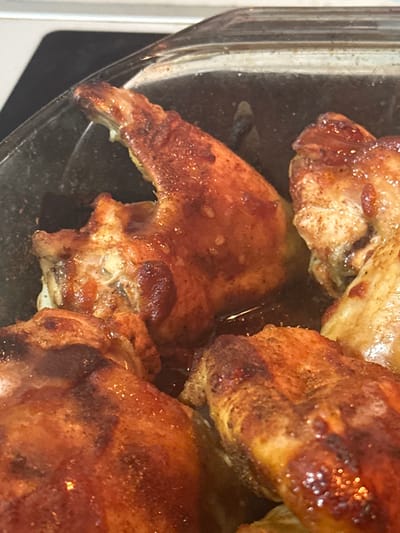The image displays a close-up, overhead view of several pieces of cooked chicken in a clear glass baking dish. The chicken pieces appear to be wings or drumettes, golden brown and glistening with a rich, dark sauce or pan juices. Their skin shows a crispy texture and some areas have a slightly charred appearance, suggesting they have been roasted or baked.

The chicken is bathed in a considerable amount of glossy, dark brown liquid at the bottom of the dish. The individual pieces are arranged closely together, overlapping slightly. Details like the texture of the chicken skin and the reflections on the sauce are clearly visible due to the close-up framing.

The setting appears to be indoors, likely a kitchen, but no specific features of the room or background are discernible other than the clear glass of the baking dish. There are no people visible, and no activity beyond the static presentation of the cooked food. The lighting is bright, highlighting the sheen on the chicken and sauce. There is no visible text in the image.