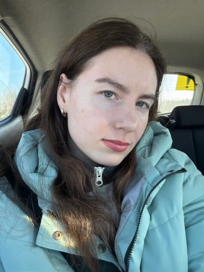 An adult female is depicted in a forward-facing photograph, seated within the passenger compartment of a vehicle in Kaykino, Russia, during daylight. The subject possesses long brown hair, blue-green eyes, light skin with visible freckles, and is wearing light pink lipstick. A small, gold-toned hoop earring is present in her left ear. She is dressed in a light mint green or teal puffer jacket over a dark grey turtleneck shirt. A silver-colored brooch, shaped like a zipper pull, is affixed to the collar of the turtleneck. The vehicle's light-colored headliner is visible above. Through the vehicle's side window, a blue sky and trees are discernible. The rear window displays distant vegetation under a bright sky and a yellow rectangular object with a black border and a black exclamation mark-like symbol adhered to its interior surface. The subject is looking directly at the camera.