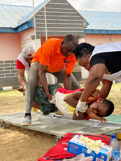 The image depicts a first aid scene. A young man in a yellow, white, and red shirt is being supported by three adults. One person, wearing a white and black top, supports his upper body. Another person, wearing an orange shirt, supports his torso. A third individual, wearing a white and red shirt and a white cap, supports his legs. The young man has a white bandage on his left arm. Medical supplies, including a box of Fitone examination gloves and a red first aid kit, are nearby. The scene appears to be outdoors, possibly in Nassarawa, Nigeria. A building with a blue and white roof is visible in the background.