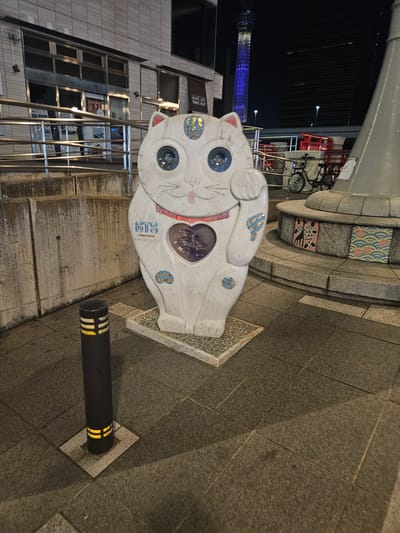 A white, sculpted cat statue with a waving right paw, decorated with blue and yellow designs, stands on a patterned base in Taito, Japan. The statue has a heart-shaped opening revealing visible objects inside. A black bollard with yellow and white stripes is anchored in the pavement nearby. The base of a structure with decorative tiles and bicycle parked behind it is visible. In the background, a building with multiple windows and a reflective metal railing is present. In the distance, a lit tower structure can be observed against the night sky. The ground consists of paved stone.