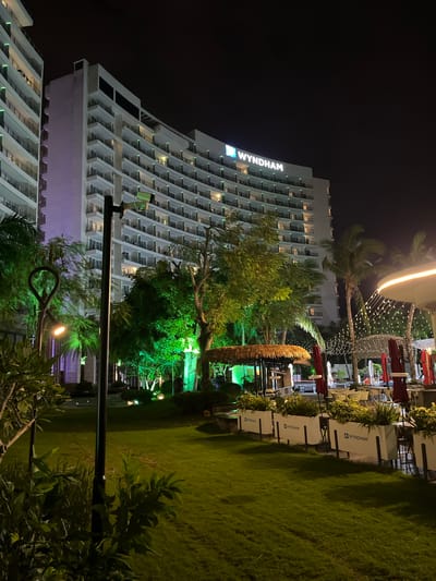 This media file depicts a nighttime scene at a resort, likely in Tianya District, China. The prominent subject is a large, modern hotel building with multiple floors and balconies, illuminated by interior lights. The "WYNDHAM" logo is clearly visible on the upper portion of the hotel.

In front of the hotel, an outdoor seating area with tables, chairs, and red umbrellas is set up on a well-maintained lawn. String lights are draped across the area, creating a festive ambiance. Palm trees and other lush greenery are scattered throughout the scene, some of which are illuminated with green lights. The overall setting suggests a resort or a luxury hotel with outdoor recreational facilities.

The time of day is clearly nighttime, indicated by the dark sky and artificial lighting. The weather appears clear, with no visible signs of rain or wind. The atmosphere is tranquil and inviting, suggesting a relaxing evening at the resort. There are no people visible in the image, focusing the attention on the architecture and the landscape. The scene is well-lit, highlighting the modern architecture of the hotel and the tropical vegetation. The presence of Wyndham branding suggests this is likely a hotel associated with that chain.