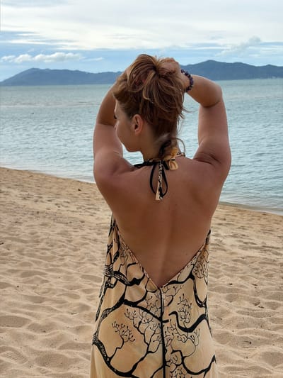 A woman is standing on a sandy beach in Ko Samui District, Thailand. She is facing away from the camera, with her back to the viewer. Her arms are raised, and her hands are behind her head, adjusting her hair. She is wearing a light-colored dress with a black, tree-like pattern. The dress has a low back with black ties at the neck. In the background, the ocean stretches to the horizon, with a range of hills or mountains visible in the distance. The sky is overcast with clouds. The sand is a light tan color, and appears to be mostly smooth with some ripples.