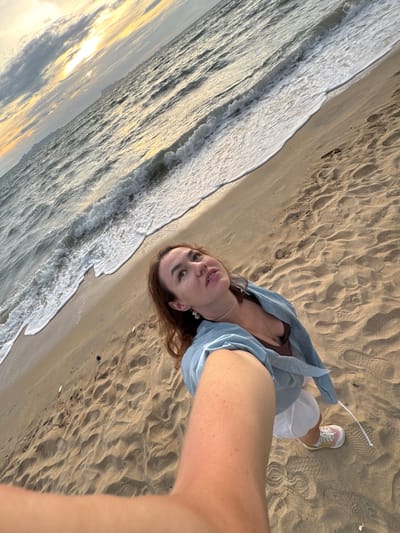 A woman is taking a selfie on a beach at sunset in Tong Yang, Thailand. The woman has brown hair and is wearing a light blue shirt over a brown top, white shorts, and sneakers. The ocean is on her left with waves breaking on the shore. The sand is golden brown and has footprints on it. The sky is a mix of orange, yellow, and gray clouds.