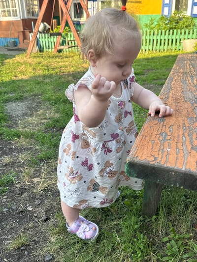 A young Caucasian female child, estimated to be between one and two years old, stands outdoors in what appears to be a rural or suburban setting in Novoyeniseyka, Russia. The child is wearing a white sleeveless dress with a repeating pattern of teddy bears and bows, and light purple open-toed sandals with bows. The child has fair hair tied up with an orange hair tie in a small ponytail. Her gaze is directed downwards and to her right, with her right hand slightly raised as if interacting with something. She is positioned next to a weathered wooden bench, part of which is visible to the right of the frame, its surface showing signs of peeling orange paint. The ground around the child is a mix of grass, dirt, and small stones. In the background, there is a wooden structure resembling a swing set or play equipment, a green wooden fence, and a building with colorful painted walls, consistent with rural Russian architecture. The lighting suggests it is daytime, with sunlight casting shadows.