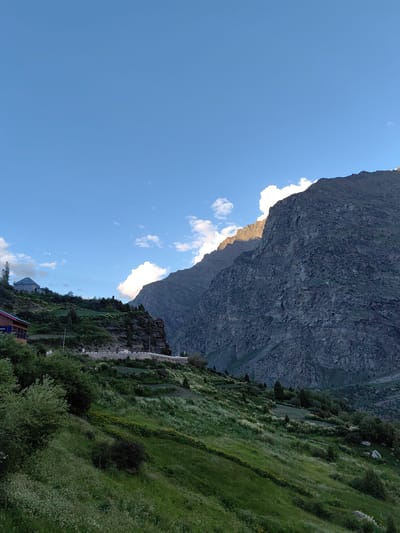 This media file captures a scenic mountain landscape in Kead, India, bathed in the golden light of late afternoon. The scene is dominated by a towering, rugged mountain on the right, its rocky face partially illuminated by the setting sun, casting dramatic shadows. In the midground, a verdant, terraced hillside slopes gently upwards, dotted with scattered trees and a carpet of wildflowers. A road winds along the base of the hillside, with a few cars visible, suggesting a rural or semi-remote location.
Perched on the higher slopes of the hill, a traditional Indian house with a distinctive architectural style is visible, its roof a deep blue. To its left, another smaller building with a red roof and wooden accents is partially obscured by foliage. The sky above is a clear, bright blue, with a few wispy clouds near the horizon, particularly around the peak of the mountain, hinting at fair weather. The overall impression is one of serene natural beauty and peaceful rural life. The time of day is clearly indicated by the low angle of the sunlight, which creates a warm, golden glow and emphasizes the textures of the landscape. There is no visible text in the image.