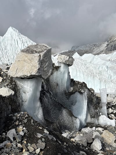 This media file captures a stark, high-altitude glacial landscape, likely in the Khumjung region of Nepal. The scene is dominated by massive formations of ice and rock under an overcast sky.
In the background, to the left, are towering, snow-covered mountain slopes partially obscured by thick, gray clouds, suggesting a cold and misty environment. The mid-ground features a large expanse of white and light blue glacier ice, characterized by textured, wavy patterns on its surface, indicating movement or melt-freeze cycles.
Towards the foreground and right, the terrain transitions into a rugged mix of dark, jagged rocks and glacial debris (moraine), interspersed with pockets of ice. A prominent, large, light-colored boulder sits in the middle-left foreground, casting a shadow and contrasting with the darker ground. Further right, the ground is covered extensively with various sizes of gray and dark rocks and gravel.
Stepped or tiered sections of dirty ice and rock appear in the center-right, possibly forming part of a glacial meltwater channel or an icefall. No people, human structures, or visible text are present in the image. The overall impression is one of a remote, powerful, and inhospitable natural environment.