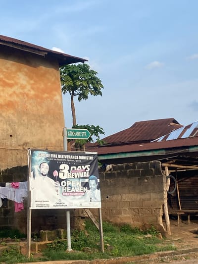This image captures a street scene in Igarra, Nigeria, specifically near the Guest Inn Road Uffa Quaters. In the foreground, a large white banner advertising a religious event is prominently displayed. The banner, from "GOD OF FIRE DELIVERANCE MINISTRY," announces a "3 DAYS POWER PACKED REVIVAL" with the theme "OPEN HEAVEN" and "GOD OF PRESERVATION." It features an image of "PROPHET DANIEL" holding a microphone and another man. Contact information "FOR ENQUIRY CALL: 07060833172," "CHURCH AUDITORIUM," and "TIME 5:00 PM" are visible at the bottom. To the left of the banner, a clothesline is strung, from which several items of laundry, including white and pink garments, are hanging.

Behind the banner, a weathered, ochre-colored building with a dark brown corrugated iron roof stands on the left. A green street sign, pointing right, is affixed to a pole directly behind the banner, clearly displaying "ATIKHAME STR." A tall papaya tree with green foliage is visible between the building on the left and the background structures. In the mid-ground and background, several other buildings with reddish-brown and silver corrugated iron roofs are visible under a light blue sky with scattered white clouds. A partially constructed or deteriorated concrete block wall runs across the lower part of the scene, with green grass growing in front and behind it. To the far right, a rustic wooden structure with a dark tire hanging from it is present. The overall impression is a daytime scene in a residential or semi-commercial area.