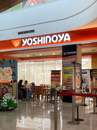 The image captures the interior of a Yoshinoya restaurant, likely located in Kudus, Indonesia, given the prevalence of Yoshinoya franchises in that region. A large, illuminated Yoshinoya logo with an orange background is prominently displayed above the entrance. To the left, a colorful children's play area features a green dinosaur ride.
Inside, three individuals are seated at a table, appearing to be a family, with two adults and one child. They are engaged in conversation or dining. The restaurant interior features wooden tables and chairs, and the floor is tiled and reflective, suggesting a well-maintained space.
Several promotional banners are displayed in the foreground, advertising various Yoshinoya deals and offers, including "GOL DEAL" and birthday party facilities. The scene is brightly lit, likely by overhead lighting within the mall or building where the restaurant is situated. The overall impression is that of a casual dining environment within a commercial space.