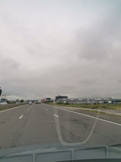 The image captures a view from inside a vehicle traveling on a highway, likely approaching or within the city of Krivina, Bulgaria. The sky is overcast with dense, grey clouds. The highway has multiple lanes, with a guardrail to the left. Several vehicles, including cars and trucks, are visible on the road. To the right of the highway, there is a large industrial building or warehouse with a sign that appears to read "WHITEFIELD PARK". Numerous trucks are parked in front of the building, and there is a patch of overgrown green vegetation between the road and the building. A construction crane is visible in the distance on the left side of the image. The road surface shows signs of wear and tear, with cracks and patches. The lower portion of the image is obscured by the dashboard and windshield of the vehicle.