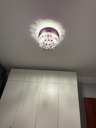 The image shows a close-up, low-angle view of a chandelier with a purple fabric shade and dangling crystals, casting a soft glow on a white ceiling. Below the ceiling, a large, modern, white wardrobe with multiple doors dominates the lower portion of the frame. A sliver of a dark curtain is visible on the left side, and on the right, a portion of a dark red door and a gray wall can be seen. The scene appears to be an indoor setting, likely a bedroom or living room, in a residential property. No people or actions are depicted. The location of Ramenskoye, Russia is not discernible from the visual information in the image.