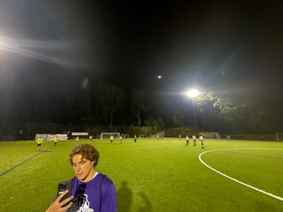 The image displays an artificial turf sports field at night, illuminated by multiple stadium lights and a visible crescent moon in a dark sky. In the foreground, a young male with wavy brown hair, wearing a purple long-sleeved shirt displaying "SPORTS" branding, holds a dark smartphone in his left hand, looking at its screen. In the midground, approximately 10-15 individuals are distributed across the green field, some wearing bright yellow/lime green pinnies and others wearing white shirts, engaged in what appears to be a soccer practice or game. Two white soccer goals are visible in the background. The field is bordered by a dark treeline and a fence. The specific location within Newton, United States, is not discernible from the image.