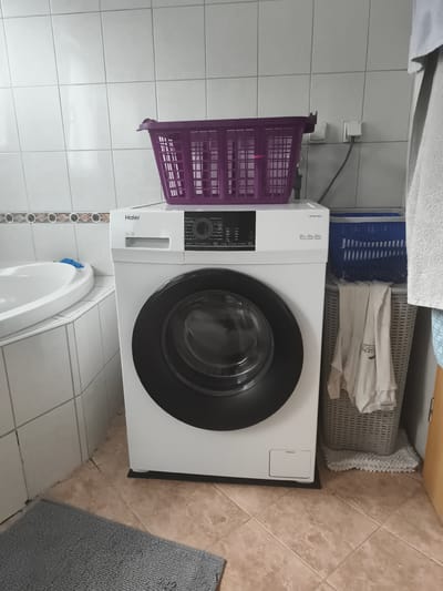 The image depicts a compact laundry area in a domestic bathroom, likely located in Eitorf, Germany. A white Haier washing machine with a black door is centrally positioned. A purple laundry basket rests on top of the washing machine. To the right of the washing machine, a blue laundry basket contains folded white clothes. Beneath this, a grey textured laundry basket holds more white clothing, one piece of which has text visible that appears to be "GEN". A grey bath mat is on the floor in the foreground. The walls are tiled in white, and the floor is tiled with a beige marble-effect pattern. The scene appears to be an indoor, residential setting, a bathroom, with no people present.