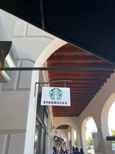 The image captures a view of a Starbucks store entrance in Rodengo, Italy. The Starbucks logo, a white rectangular sign with the iconic green siren, is prominently displayed and hung from a metal support. The store is situated beneath a series of arched walkways with exposed wooden beams. In the background, several people are visible walking towards the store and other shops, suggesting a public area or shopping center. The architecture of the building features white stucco walls and decorative elements. Further down the walkway, other signs are visible, including one that reads "Rossignol" and another with "Tax Free". The overall scene depicts a commercial area on a bright day.