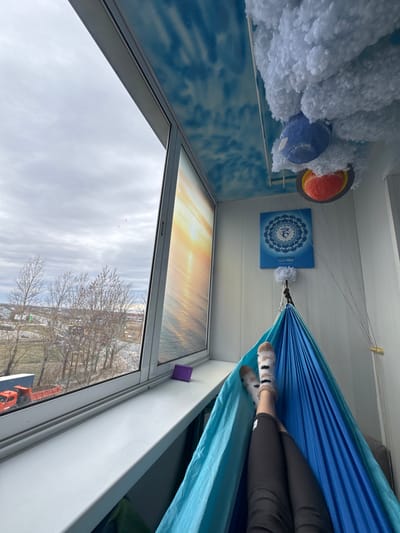 A view from inside a room looking outward through a window on what appears to be a cloudy day in Kladovitsy, Russia. Inside, a blue hammock is suspended, and a person wearing patterned socks and dark pants is reclined within it. On the wall, there's a blue poster with a mandala-like design and an orange and blue decorative item surrounded by white fluffy material, possibly representing a planet or celestial body. The room has white walls, and the window reflects a sunset or sunrise scene, which might be a painting or poster. Outside the window, bare trees and buildings characteristic of a city environment are visible. A crane is visible below the trees.