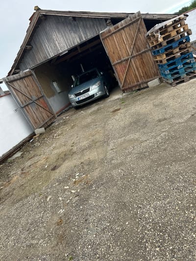 The image captures an overhead view of a grey sedan, partially parked inside a rustic, weathered wooden garage or barn. The vehicle's front end, including its headlights, grille, and part of the hood, is visible. The license plate "CC 2190 AX" is clearly legible on the front of the car, indicating an Italian registration, despite the provided location context of Dobrotitsa, Bulgaria.

The garage features large wooden double doors, one of which is open, revealing the dark interior where the car is situated. The interior floor appears to be concrete or paved, with some damp patches. Above the car, the wooden planking of the garage's ceiling or upper structure is visible.

To the left of the garage entrance, a stack of various pallets can be seen, a mix of wooden and blue plastic types. The ground surface outside the garage is a rough, granular mix of gravel and dirt, with sparse patches of green growth. The lighting is diffused, suggesting an overcast daytime, and there are no strong shadows. No people or specific activities are depicted; the scene shows a parked car in a static, outdoor setting.