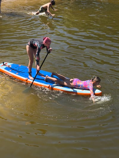 In Malakhovka, Russia, on a sunny day, two individuals are engaged in water activities. A girl with pink hair, standing on a blue and orange paddleboard, uses a paddle to navigate the murky, rippling water. Another person, lying prone on the same paddleboard, wears a colorful long-sleeved swimsuit and is partially submerged in the water, with hands reaching forward. In the background, a shirtless boy wearing patterned swim trunks is in the process of entering the water, splashing with his arms and legs. The environment suggests a natural body of water, possibly a river or lake, with visible sunlight reflecting off the surface and creating subtle patterns of light and shadow.