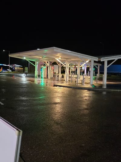The image depicts an outdoor commercial or service area at night, characterized by a brightly lit, modern canopy structure and a wet, reflective ground surface. The scene is devoid of people.
The main subject is a large, white, angular canopy, likely constructed from metal beams and a translucent roof, extending over a paved area. It is illuminated from within by warm yellow-white lights and from below by green and yellow accent lights, which cast reflections onto the wet ground. Underneath the canopy, various pieces of equipment, possibly pumps or service bays, are visible but indistinct. Towards the back of the sheltered area, parts of a building facade with additional lighting can be seen. On the left side of the frame, the dark expanse of the night sky is visible above the canopy.
The ground is a dark, wet asphalt or concrete surface, reflecting the bright lights of the canopy structure. The wetness suggests recent rainfall or ongoing precipitation. These reflections create streaks of light, especially green and yellow, across the dark pavement.
Notable details include the time of day being night, evident from the dark sky and reliance on artificial lighting. The wet ground indicates either recent rain or a damp environment. In the bottom right corner, a small section of a white board or sign is partially visible. Within the main structure, on a blurry banner or sign, some faint text, possibly "ENTRE" (likely "Entrée" or entrance in French, given the location context of France), can be discerned. Further right, on a white vehicle, indistinct letters like "EAT" are visible on its side. The overall setting implies a functional, possibly commercial establishment such as a gas station, car wash, or drive-through service point in Longeville-lès-Saint-Avold, France.
