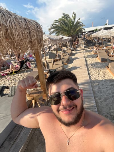 The image captures a man posing for a selfie on a sunny beach in Nea Kallikratia, Greece. The man, who is shirtless and wearing sunglasses and a gold cross necklace, flexes his bicep towards the camera. Behind him, a wooden walkway stretches along a beach lined with rows of lounge chairs and thatched umbrellas. Several other beachgoers are visible in the background, relaxing under umbrellas or walking along the pathway. Palm trees and a building with signage indicating "CABANA C5" are also present, suggesting a resort or beach club setting. The overall atmosphere is one of leisure and vacation.
