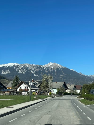 The image depicts a street in Bled, Slovenia, lined with houses and bordered by a mountain range. The sky is clear and blue. The mountains are partially snow-covered. A grey asphalt road runs along the right side of the frame, marked with white dashed lines. Houses with varying architectural styles, some with visible solar panels, are situated between the road and the mountain. Trees and green spaces fill the area around the houses.
