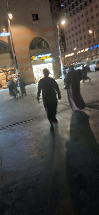 The image depicts a nighttime street scene in Medina, Saudi Arabia, characterized by artificial illumination and motion blur. The foreground features dark, reflective pavement. A central, blurred figure in dark attire walks towards the viewer, while to their right, another blurred figure in a lighter, flowing garment walks away from the camera. Both figures cast elongated shadows on the ground.

To the left midground, several indistinct individuals are present near a brightly lit commercial building. This building exhibits a light-toned facade with prominent arched ground-floor entrances. A shop sign above one arch displays yellow Arabic script, appearing to read "أسواق الشيخ" (Aswaq Al-Sheikh), alongside a distinctive yellow logo. The shop interior is brightly lit, revealing merchandise.

In the background to the right, a tall, multi-story building with numerous illuminated windows is visible, consistent with a hotel or large commercial structure. Faintly discernible vehicles are present on a street further in the background. Streetlights contribute to the overall illumination, casting light and reflections across the wet-appearing ground.