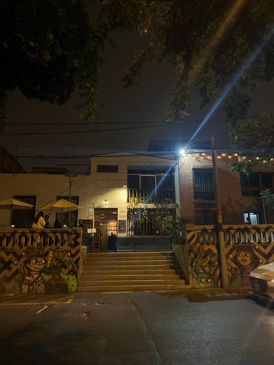 An eye-level, long shot taken at night shows a street in Barranco, Peru. The buildings lining the street are a mix of brick, concrete, and painted surfaces, lit by street lights and lights from within buildings. The nearest building has colorful, stylized mural art on the facade. A flight of stairs leads to an entrance. People sit at tables beneath open umbrellas in front of one building. Trees with dense foliage are on the left side. Electrical wires crisscross the upper portion of the frame. The street is asphalt, and appears slightly wet.