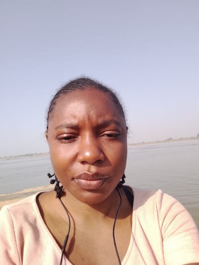 The image captures an outdoor scene in the city of Ibi, Nigeria, focusing on a single individual in the foreground. An adult female, with dark brown skin and hair styled in cornrows, faces the camera directly. Her facial expression is neutral, with slightly furrowed brows and open, subtly downcast eyes. She is wearing a light peach-colored, ribbed, short-sleeved top. Black wired earphones are visible around her neck, with the cable extending over her chest. Small, dark, decorative elements, possibly woven, are present near her ears. The background features a wide body of calm water, consistent with a river or large lake, stretching horizontally across the mid-ground. A hazy, distant shoreline with faint vegetation is discernible across the water on the left side. The sky is clear and light blue, indicating daylight. A concrete or paved structure separates the individual from the water immediately behind her.