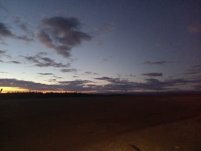 The scene depicts an expansive outdoor landscape at twilight, likely dawn or dusk, located in Santa Juana, Chile. The sky occupies the upper two-thirds of the image, transitioning from a dark blue-grey overhead to lighter blue and orange tones at the horizon, where a distinct orange glow is present. Scattered stratocumulus cloud formations are visible, with denser, darker clusters in the upper left quadrant. A continuous, dark, silhouetted line of what appears to be a coniferous forest or trees defines the horizon across the mid-ground. A thin, dark, vertical silhouette is partially visible on the far left, extending marginally above the tree line. The lower third of the image consists of a dark, flat, open ground, appearing as bare, reddish-brown earth, with no discernible features or structures. A faint, diffuse light source illuminates a small, indistinct area in the lower right foreground. No individuals, animals, or prominent man-made objects are visible within the depicted landscape.