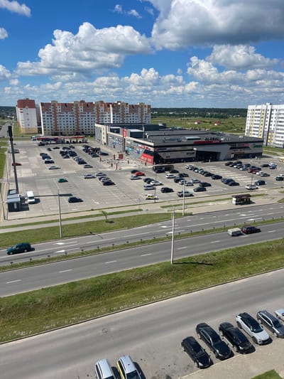 This is a high-angle, wide shot of a commercial area on a sunny day in Rechytsa, Belarus. The scene is dominated by a large, modern shopping mall named "GLOBAL market" with prominent signage. In front of the mall is an expansive, paved parking lot filled with numerous cars. To the left of the mall, several multi-story residential buildings with orange and white facades are visible. A gas station is situated between the residential buildings and the mall parking lot. A multi-lane road runs along the bottom of the frame, with a few cars driving on it. The environment includes green grass and trees interspersed with the urban structures. The sky is blue with scattered white clouds. There are no visible people in the image.