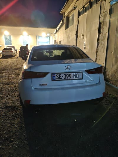 A white Lexus IS 250 is parked on a gravel surface in front of a light-colored building with large metal doors. The license plate reads "SE-099-MO GE". The car is facing away from the camera. In the background, two other vehicles are visible, one white and one dark. A person wearing a dark shirt is standing next to the dark vehicle. The building appears to be a workshop or garage. The scene is lit by artificial lights. This is likely a scene in Krtsanisi, Georgia.